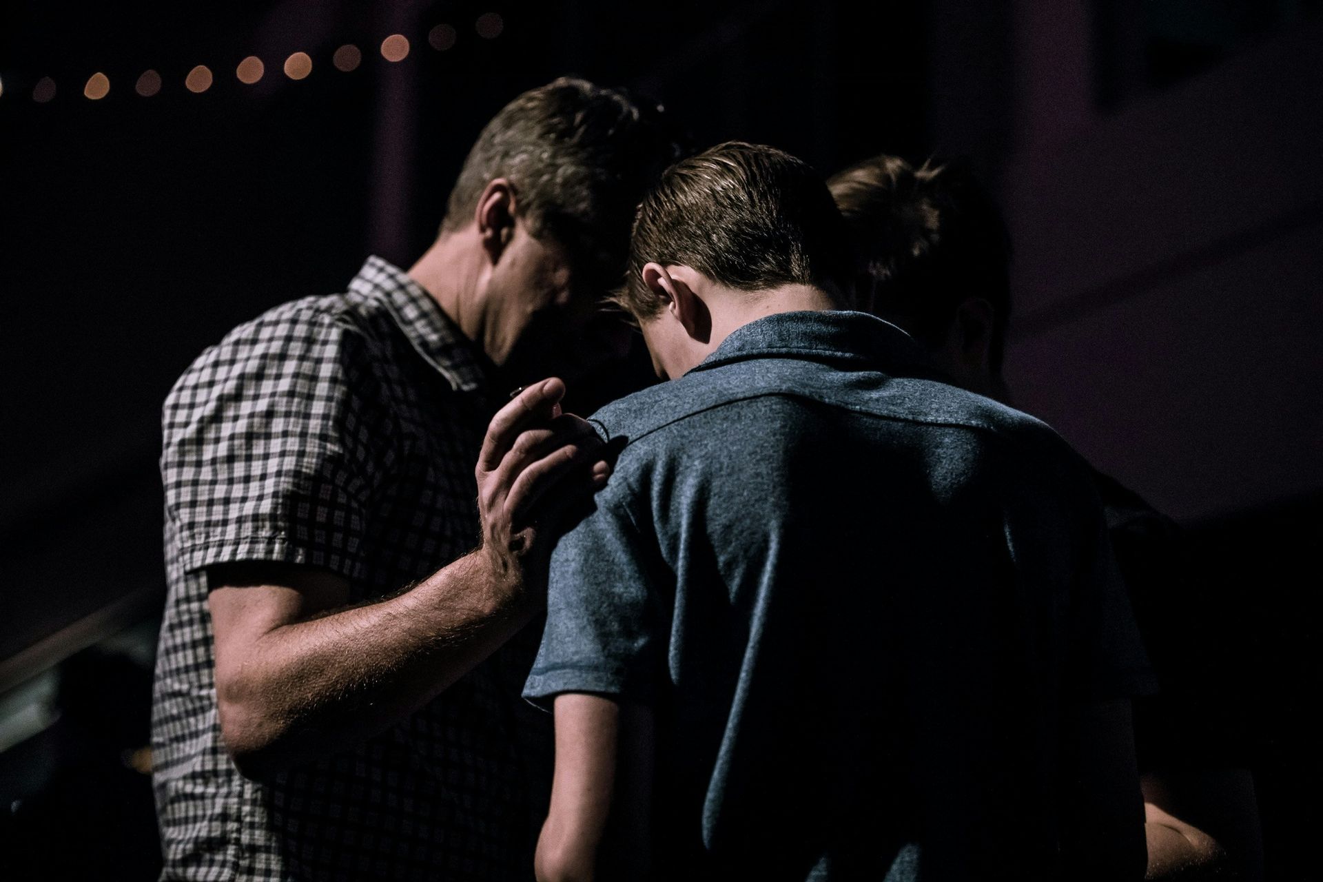 Two men standing close together in dim light, one with a hand on the other’s shoulder.