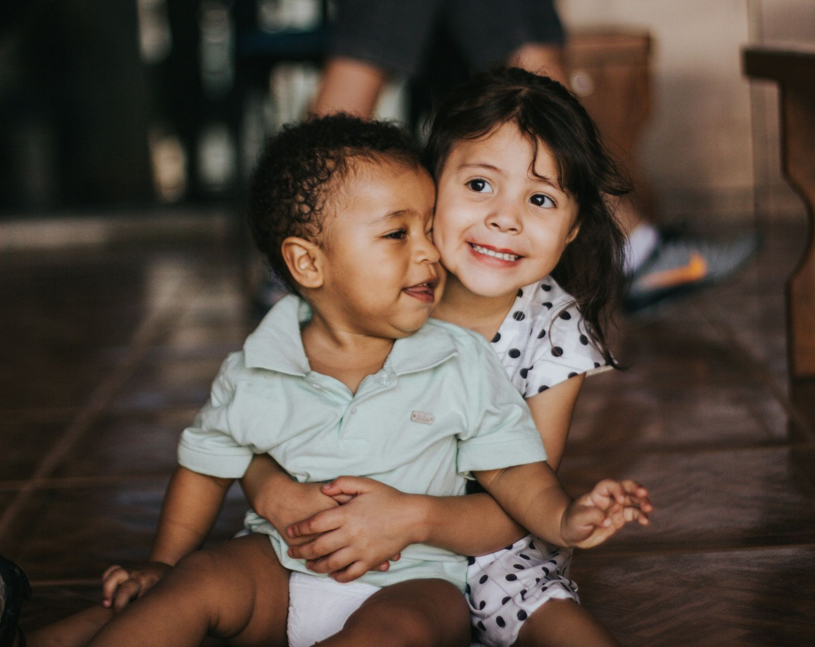 Two young children sitting close together indoors, smiling and hugging on a wooden floor.