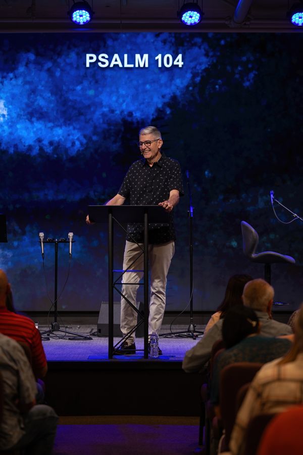 Speaker at a lectern on a church stage under blue lights, with “PSALM 104” displayed behind him.