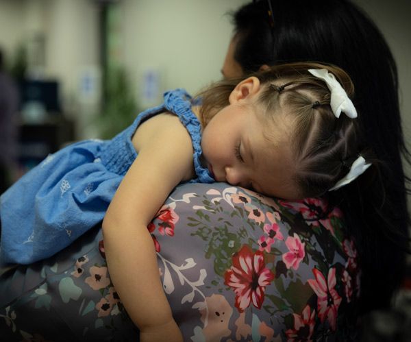 Sleeping child resting on an adult’s shoulder, wearing a blue dress with a floral floral top in a soft indoor setting