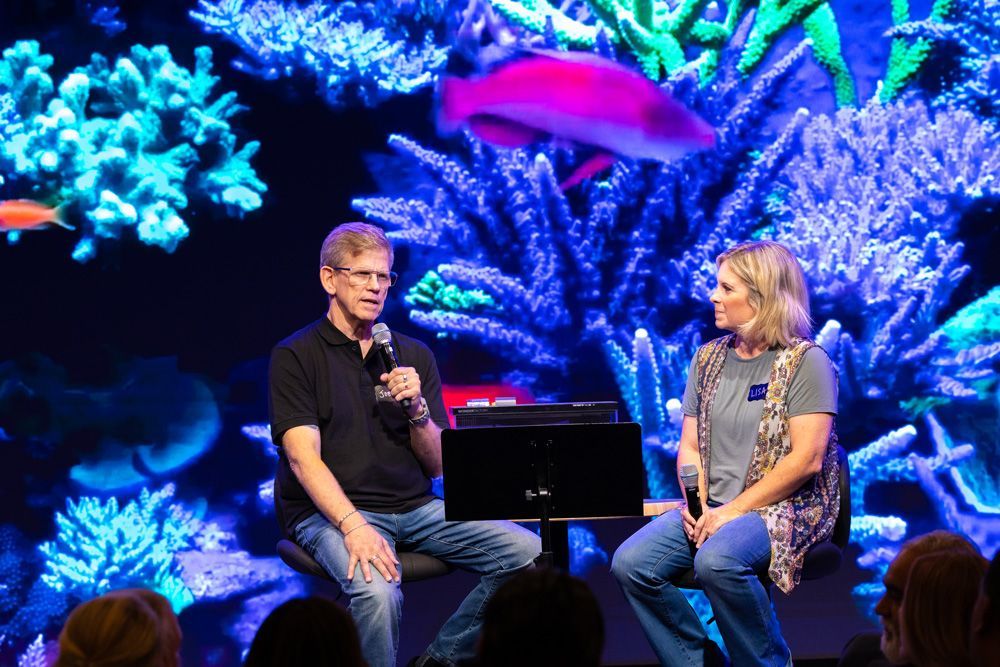 Two people sit on stage speaking into microphones before a vivid blue coral reef backdrop