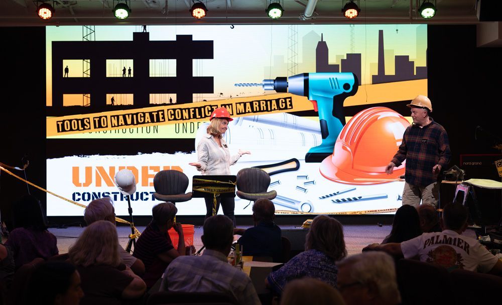 Conference stage with a giant screen showing a blue camera and orange graphics, panelists seated below.