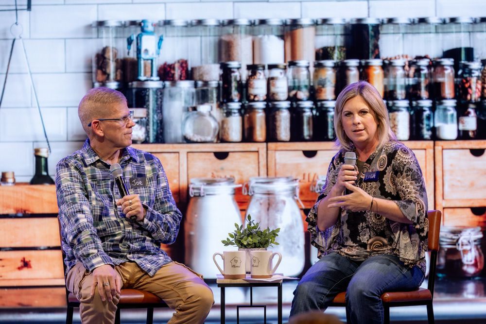 Two speakers seated on stage with microphones in front of shelves of bottles and jars