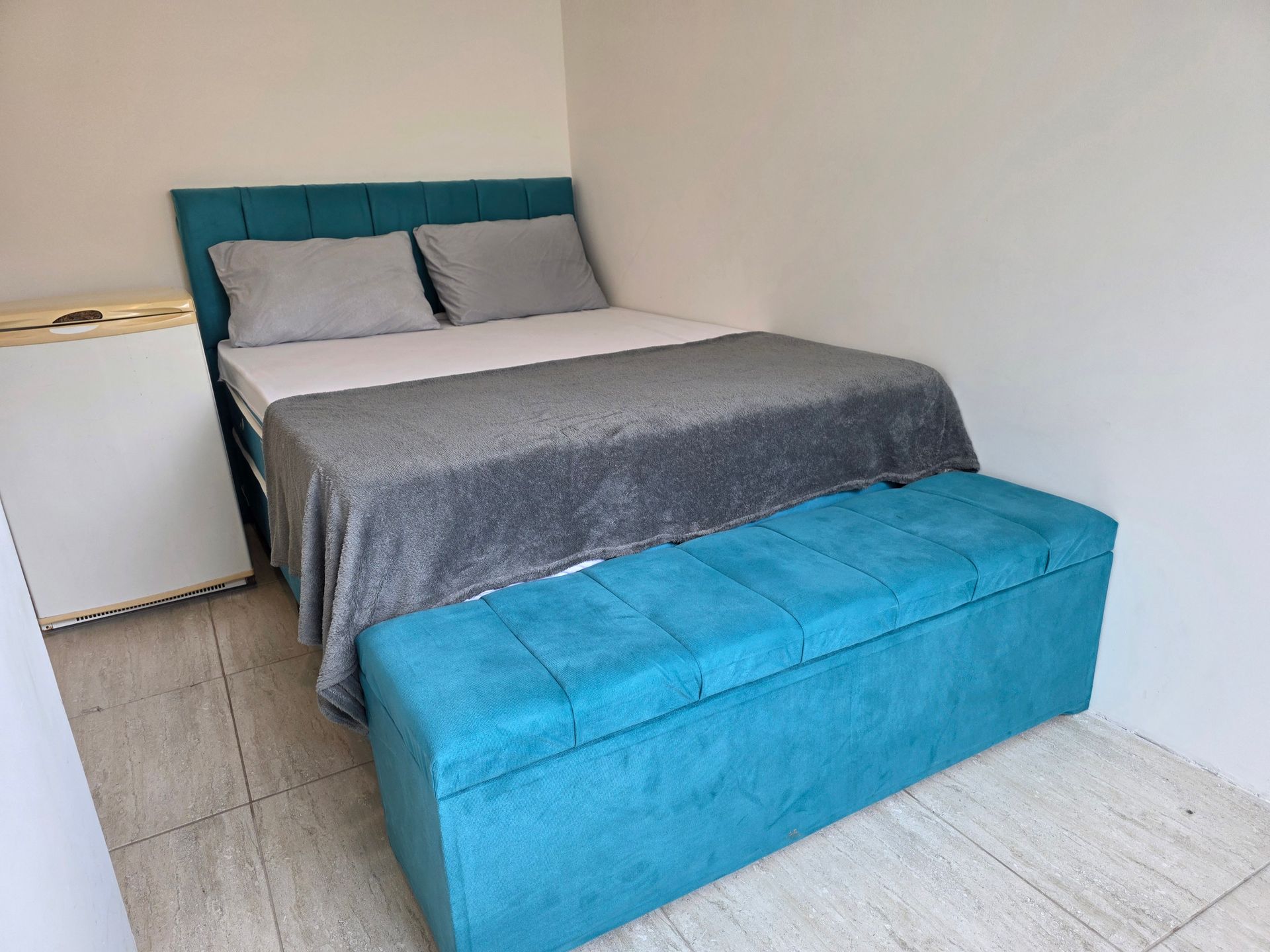 Cama estofada em tom turquesa e banco com espaço para armazenamento em um quarto pequeno. Roupa de cama cinza e um frigobar também são visíveis.