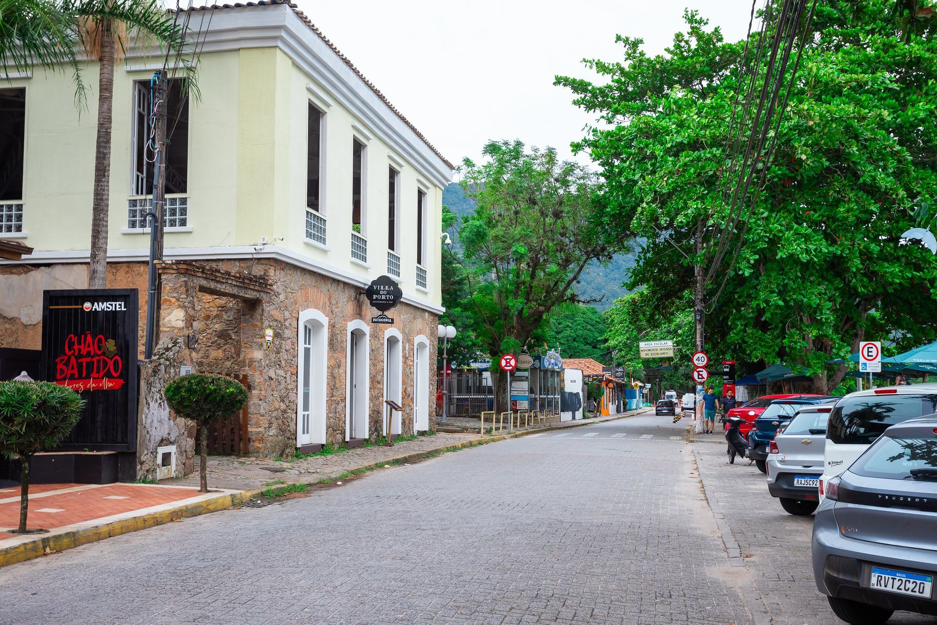 Rua de paralelepípedos ladeada por prédios e carros estacionados; árvores exuberantes margeiam a rua.