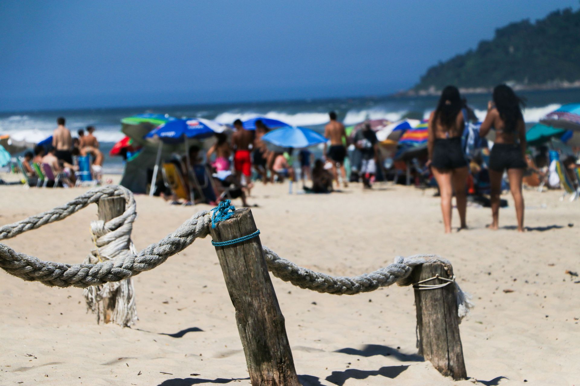 Cerca de corda em uma praia de areia, pessoas e guarda-sóis ao fundo, oceano e céu azuis.