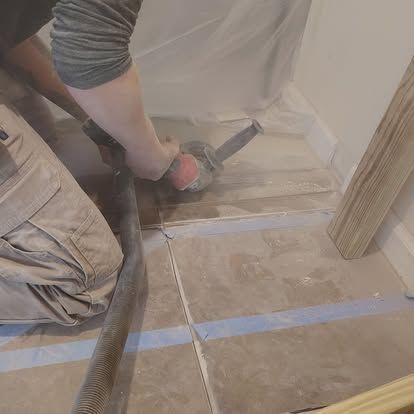 Person using a power tool to cut flooring, producing dust, indoors. A vacuum hose is attached.