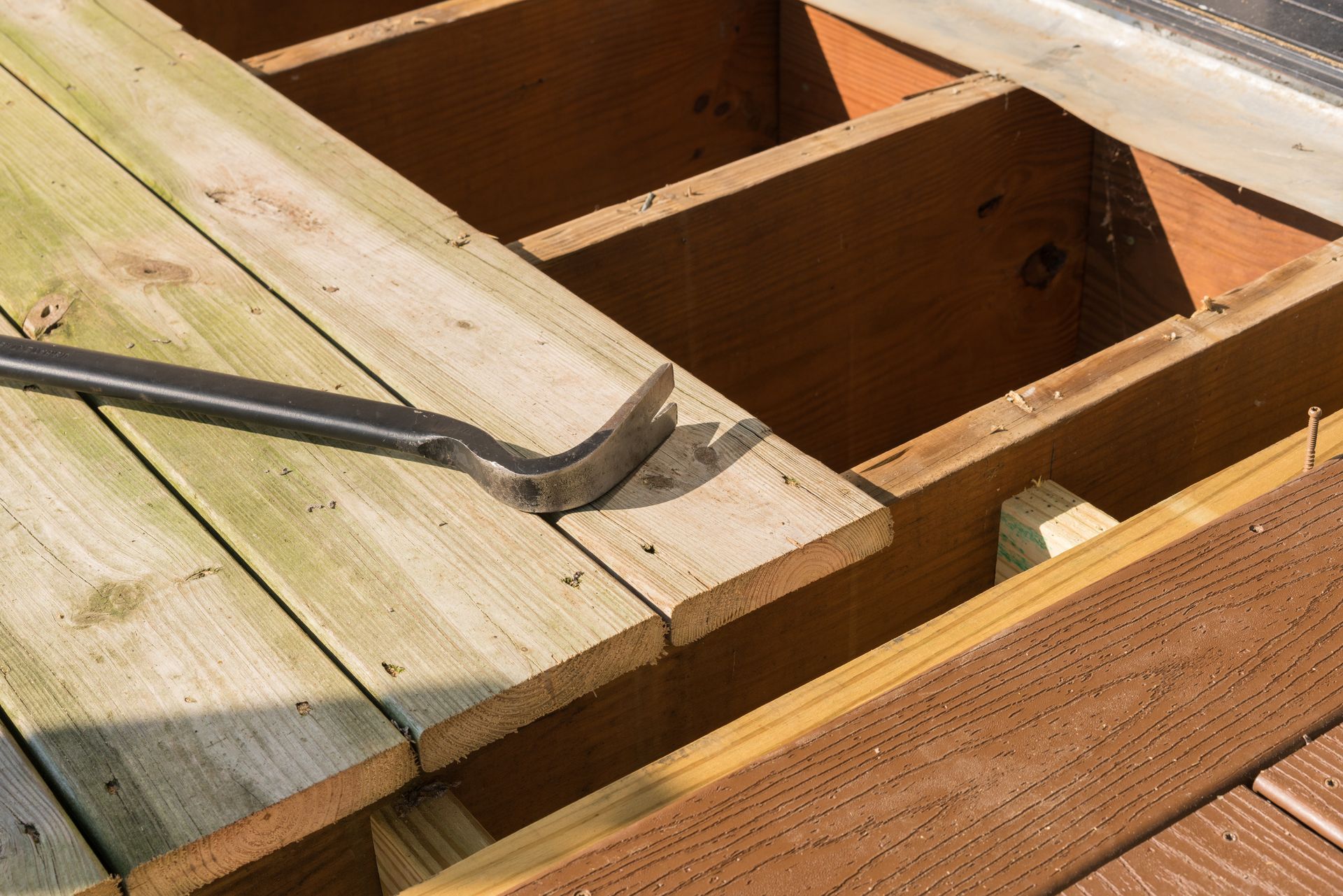 Pry bar lifting weathered wooden deck boards over exposed joists during repair work