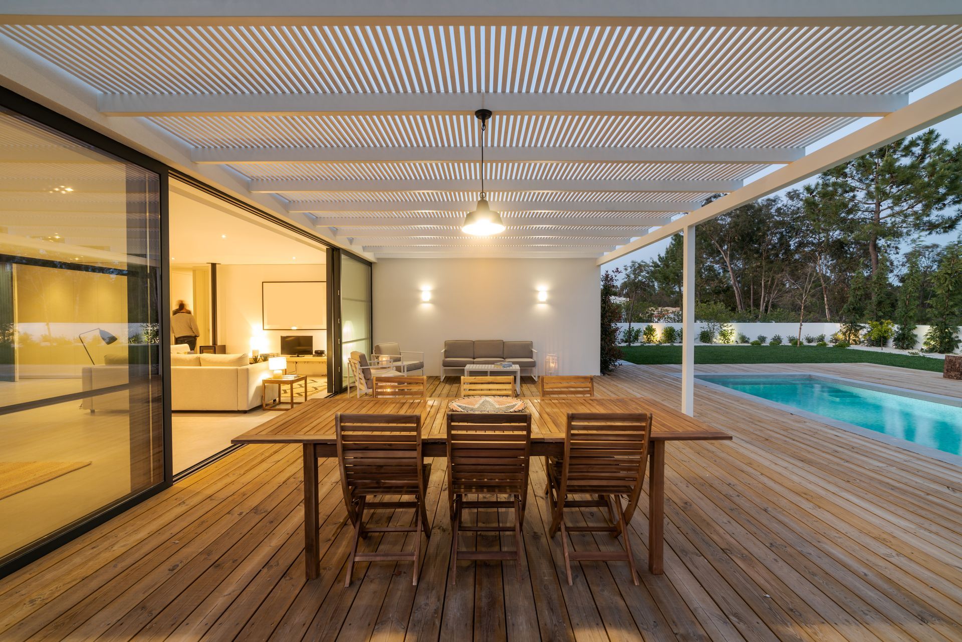 A wooden deck with a table and chairs under a pergola next to a swimming pool.