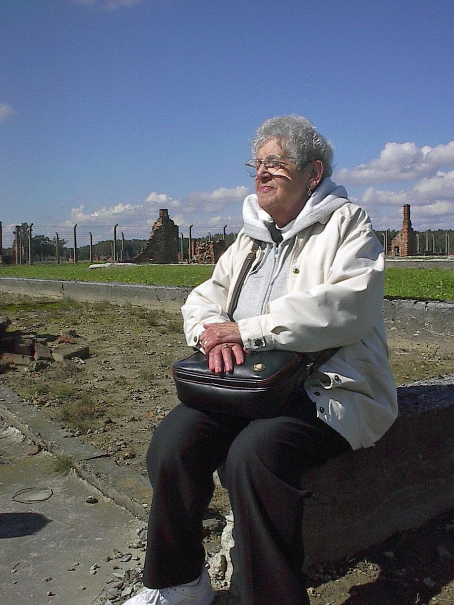Person in a white jacket sitting outdoors on a stone bench, holding a black bag, with a grassy field and sky behind.