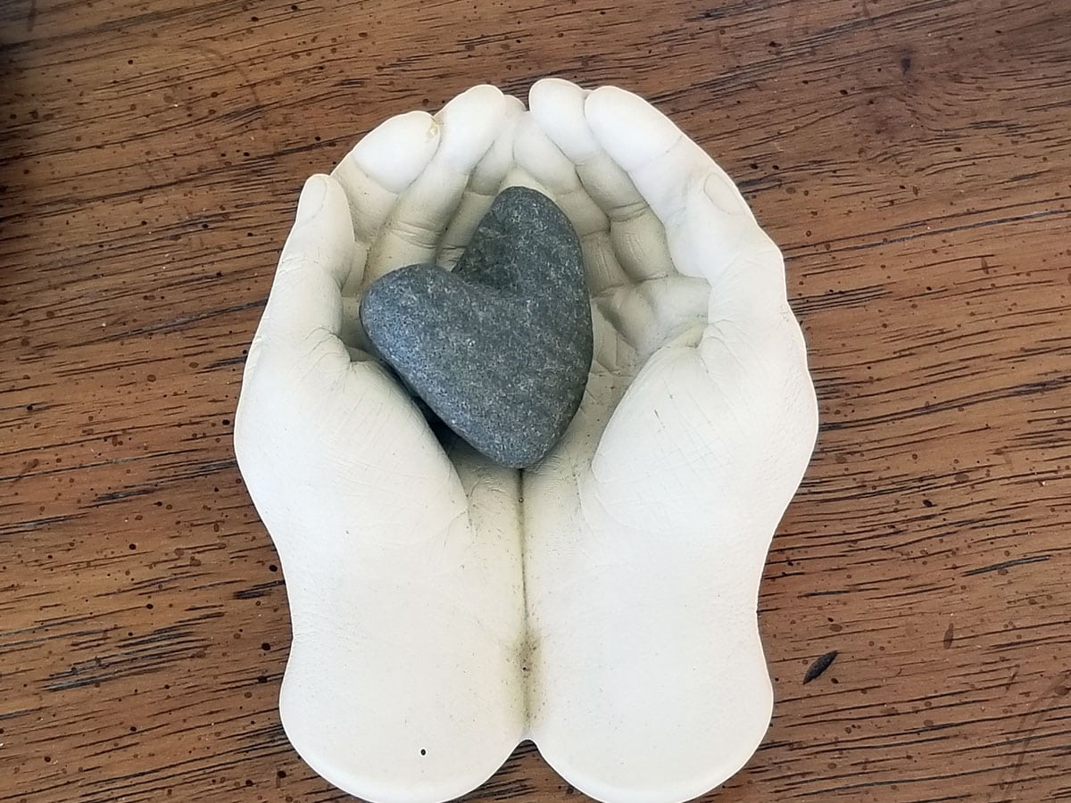 Two white cupped hands holding a gray heart-shaped stone on a wooden surface