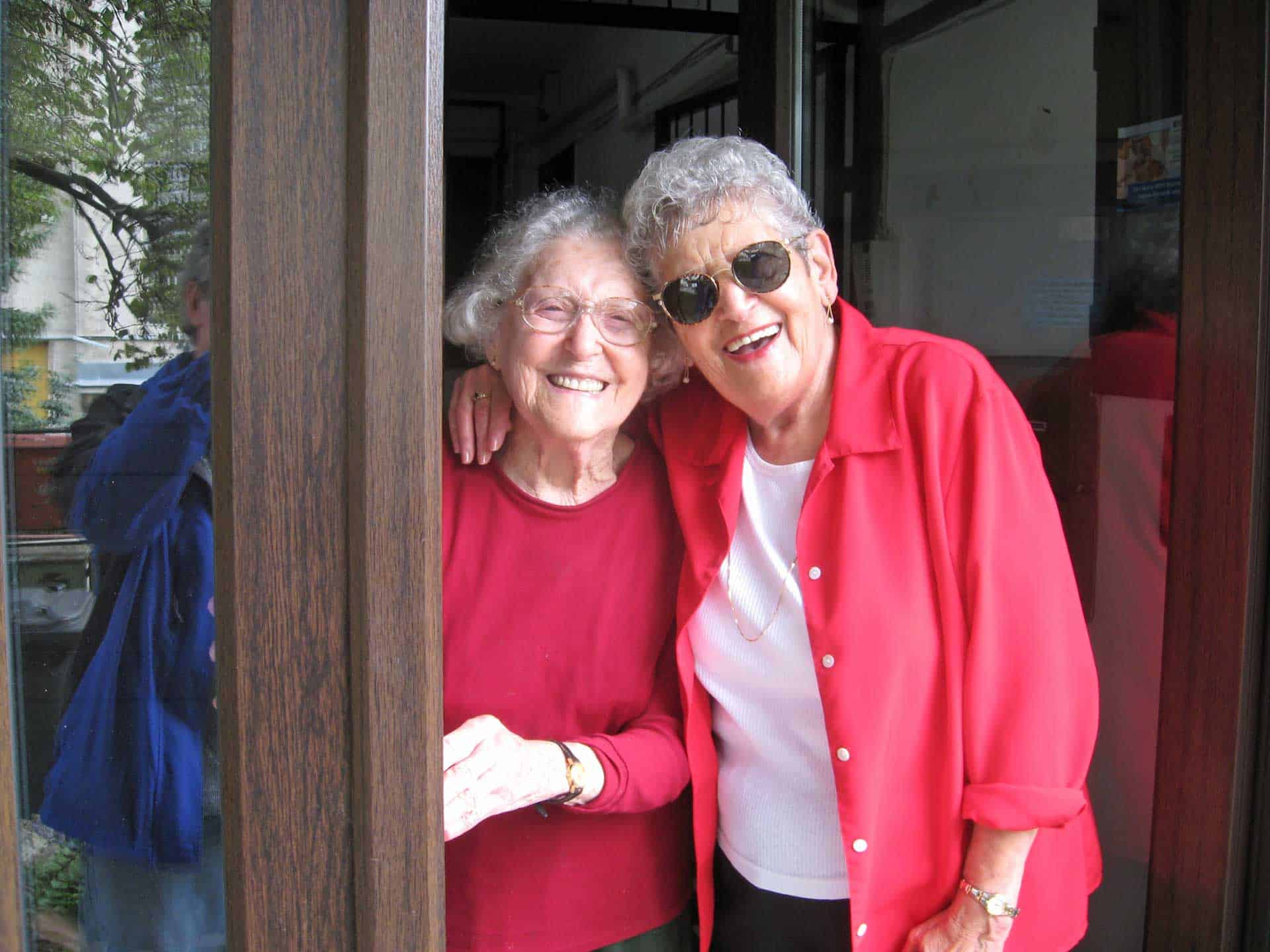 Two smiling women standing arm-in-arm at a doorway, both wearing red jackets.