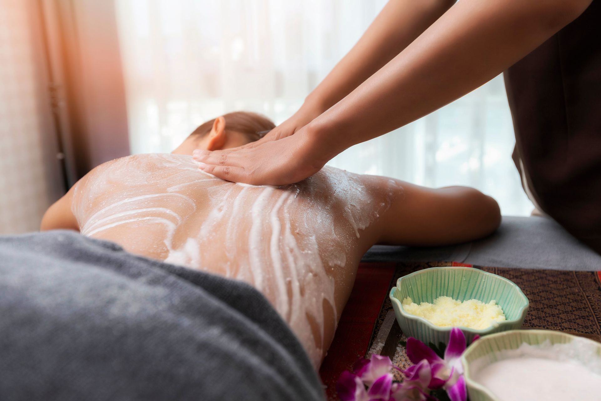 Massage therapist applying oil to a person’s back on a spa table, with towels and flower petals nearby