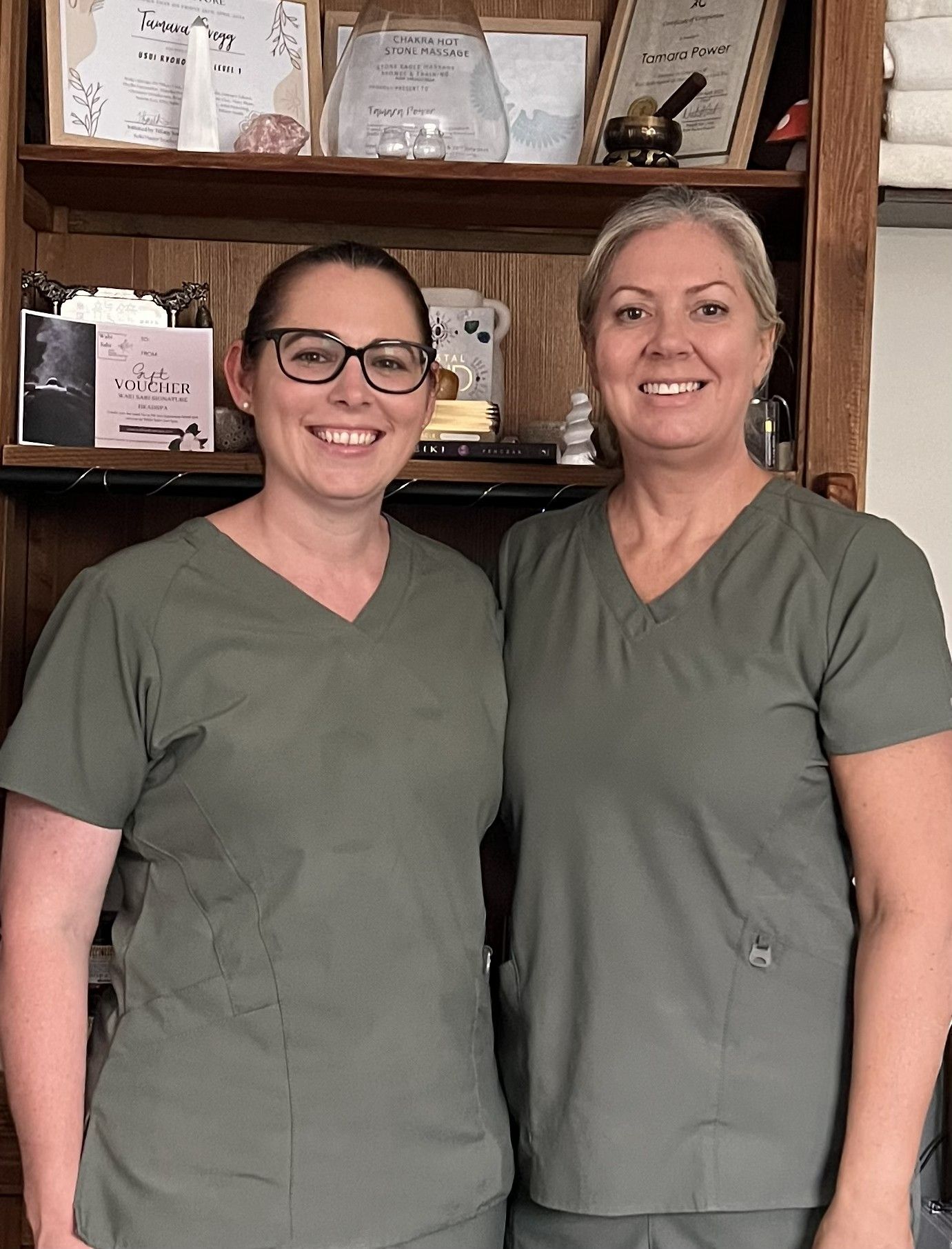 Two smiling people in green scrub tops standing in front of shelves with framed certificates.