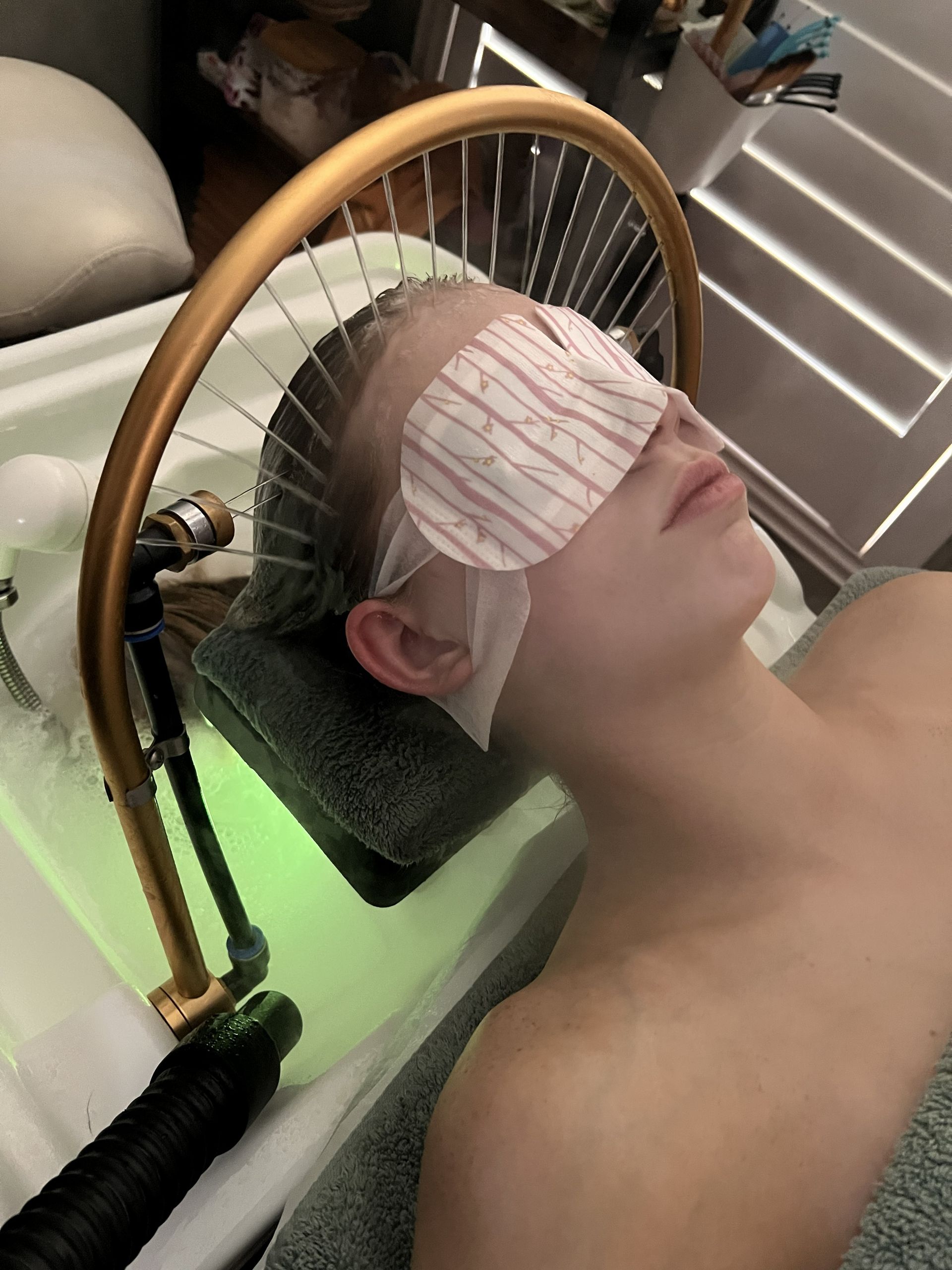 Person lying back with a pink eye mask on, resting near a wooden wheel and blinds.