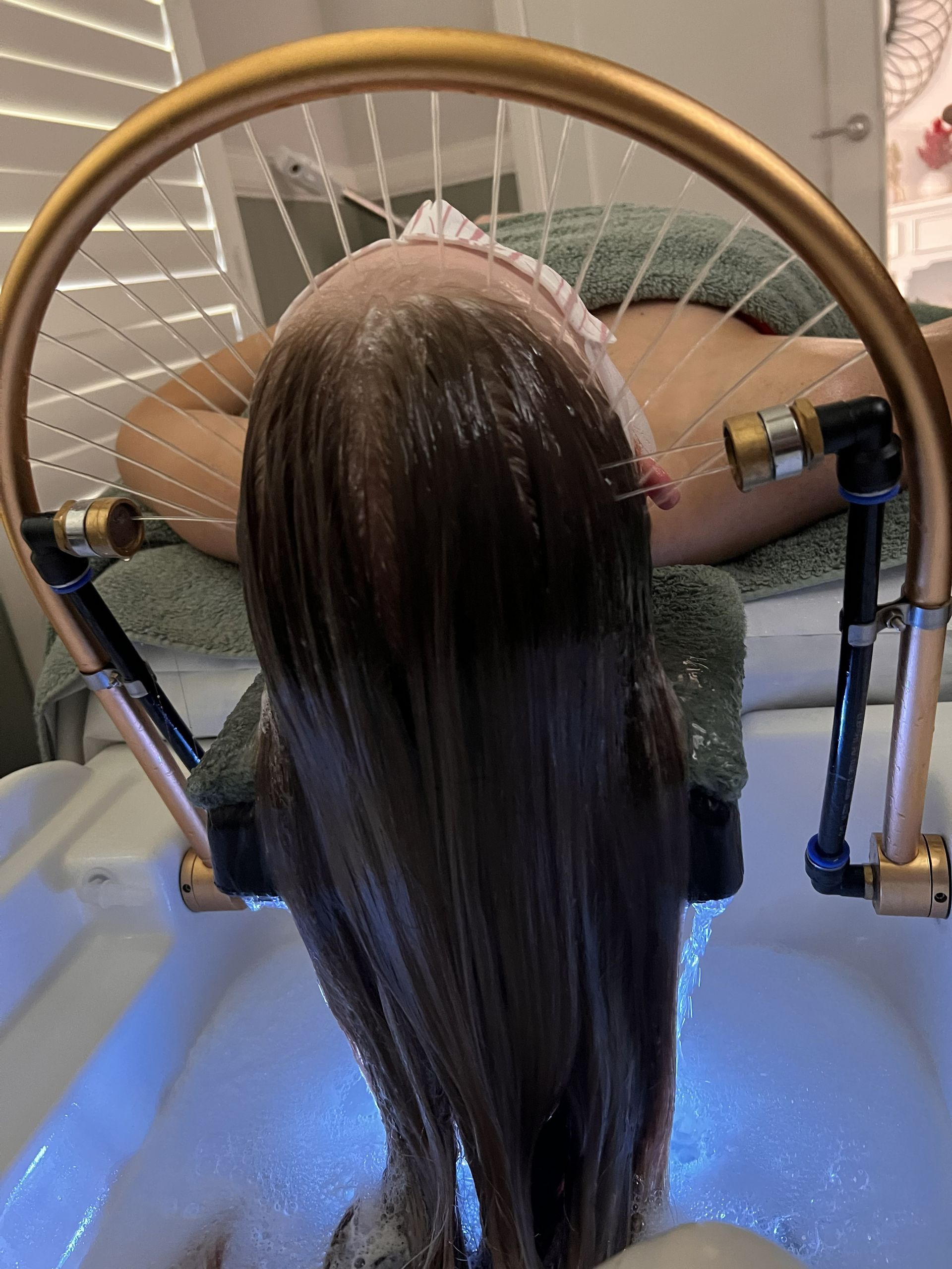 Hair being washed at a salon shampoo bowl under running water, viewed from behind.