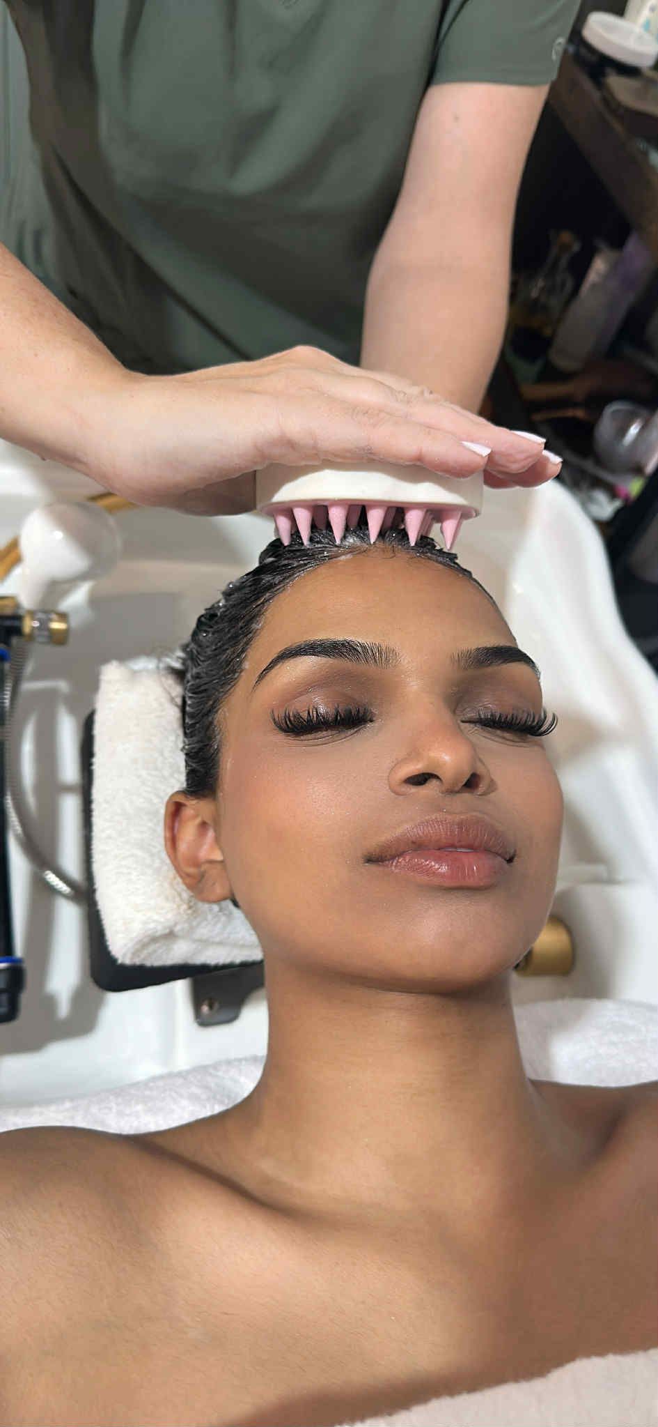 Person receiving a scalp treatment at a salon sink, eyes closed, with a towel and stylist’s hands above head