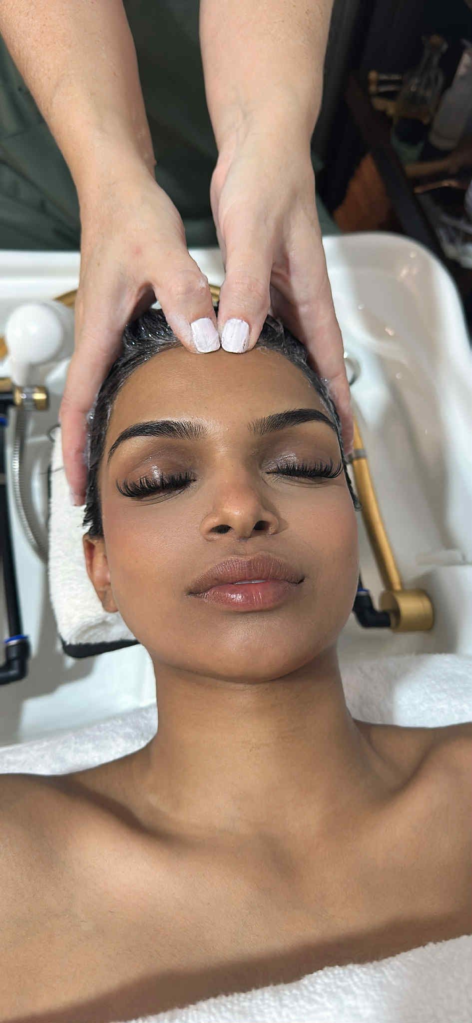 Woman receiving a facial massage at a spa sink, eyes closed, with white towels and skincare tools nearby