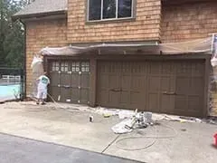 Worker painting garage doors on a house exterior with brown siding and tarps on the driveway