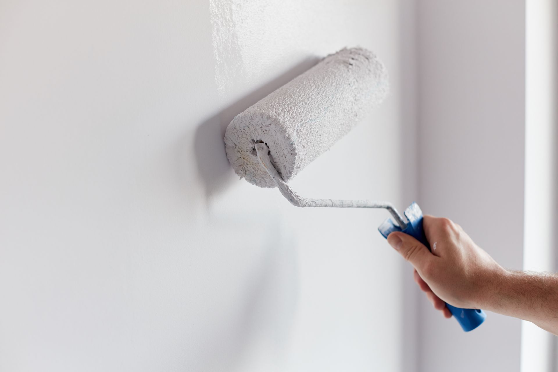 Hand using a paint roller with a blue handle to apply paint on a smooth wall.