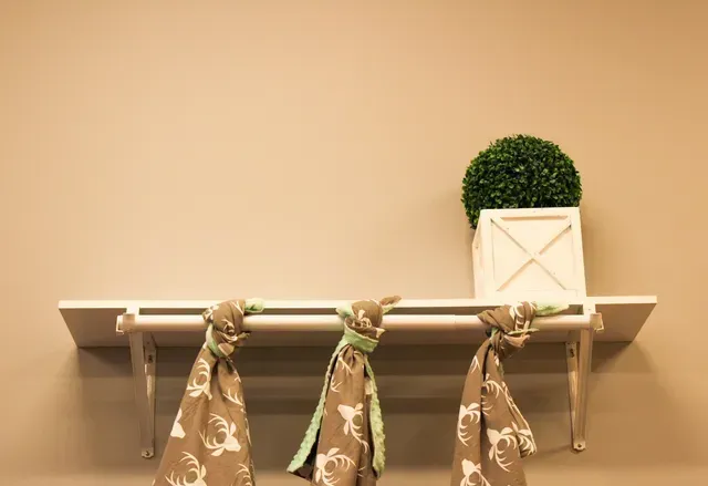 White shelf with patterned cloths hanging beneath and a small green potted plant on top against a beige wall