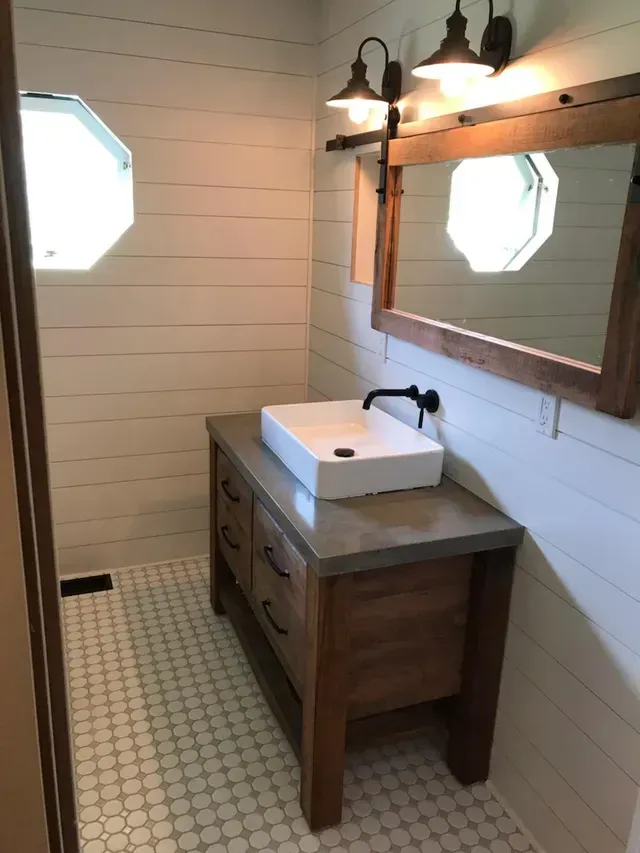Small bathroom vanity with white sink, wood cabinet, mirror, and wall sconces on tiled floor.