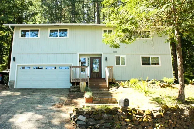 Two-story light green house with garage, front porch, and wooded yard