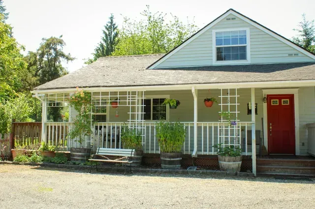 Small white house with red door, front porch, and hanging planters surrounded by greenery