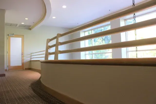Empty curved hallway with beige carpet, white railing, and large windows letting in natural light