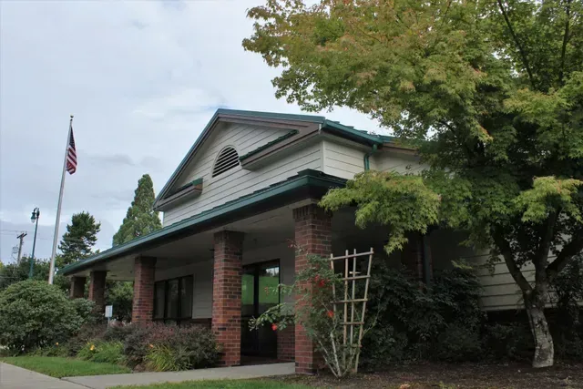 Brick building with green trim and an American flag, partly obscured by trees under a cloudy sky