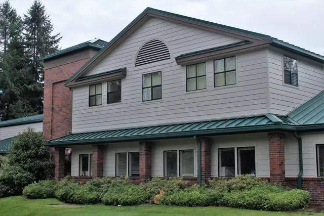 Two-story suburban house with gray siding, green roof, brick chimney, and front shrubs