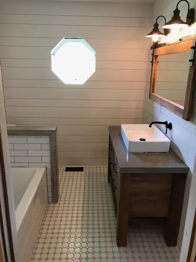 Bright bathroom with octagonal window, pedestal sink, mirror, and bathtub beside white beadboard walls.