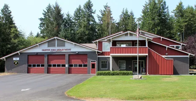 Red fire station with three garage doors and a red barn-style annex beside a grassy lawn and trees