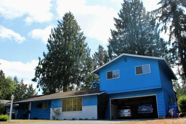 Blue two-story house with an open garage and parked car, surrounded by tall pine trees under a partly cloudy sky