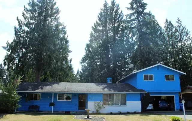 Blue house with a carport and tall evergreen trees in the yard