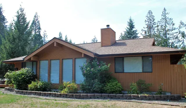 Single-story wooden house with a red roof, blue windows, and tall trees in the background