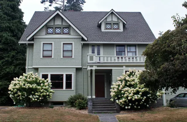 Gray two-story house with white trim, front porch, and blooming shrubs by the entrance