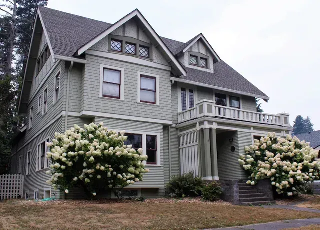 Gray Victorian house with white trim and flowering shrubs in front yard