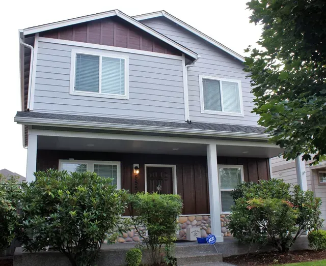 Two-story gray house with white trim, front porch, and bushes in front on a rainy day