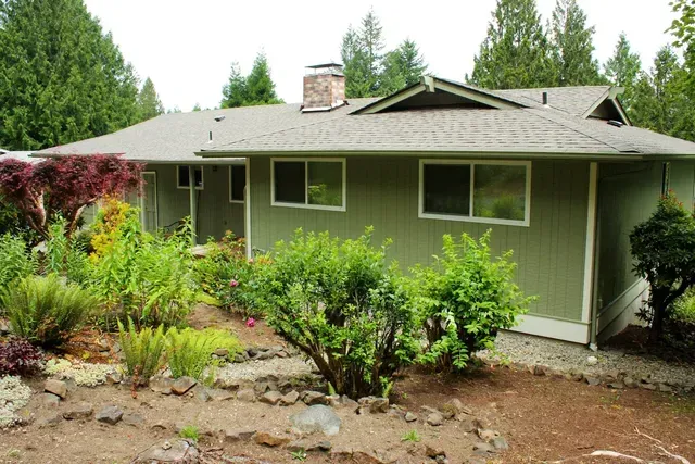 Single-story green house with a landscaped front yard and tall trees in the background