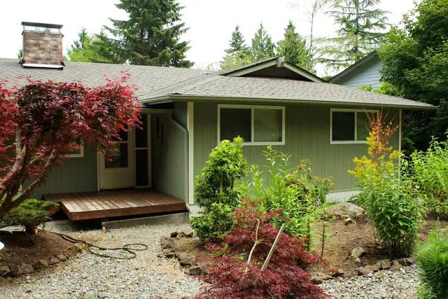 Small green house with a front porch, red and green shrubs, and a stone garden path
