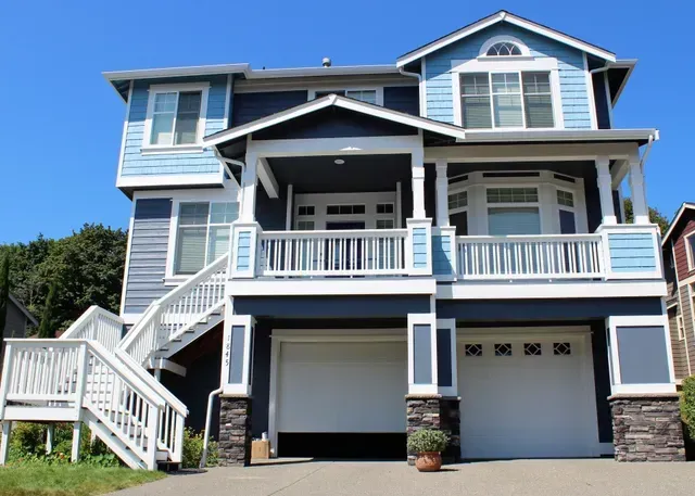 Large light-blue two-story house with white trim, front porch, staircase, and attached garage under a clear sky