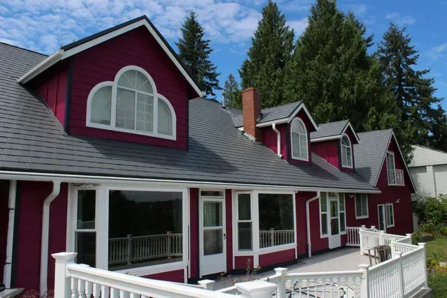 Maroon house with white trim, arched dormer windows, and a porch beside tall evergreen trees