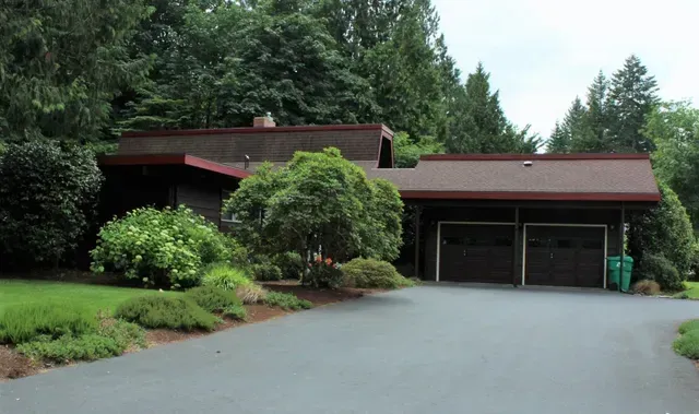 Modern house with a double garage, framed by trees and shrubs, beside a paved driveway