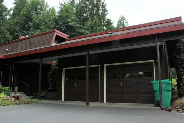 Driveway facing a house with a red roof, garage doors, and green trash bins under trees