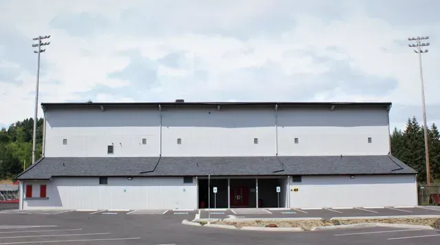 Exterior of a large white sports building with a gray roof and parking lot in front