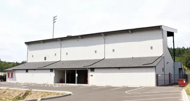 Large white sports building with gray roof, entry doors, and parking lot outside