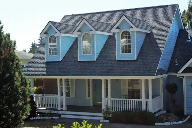 White two-story house with dormer windows, gray roof, and a covered front porch surrounded by greenery