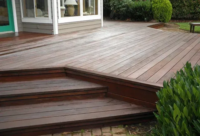 Wooden deck with steps leading to a house, bordered by green shrubs and a small lawn.