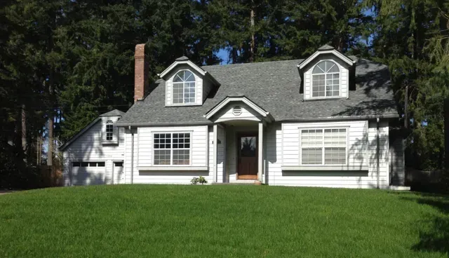 White house with gray roof and dormer windows, front porch, and large green lawn under trees